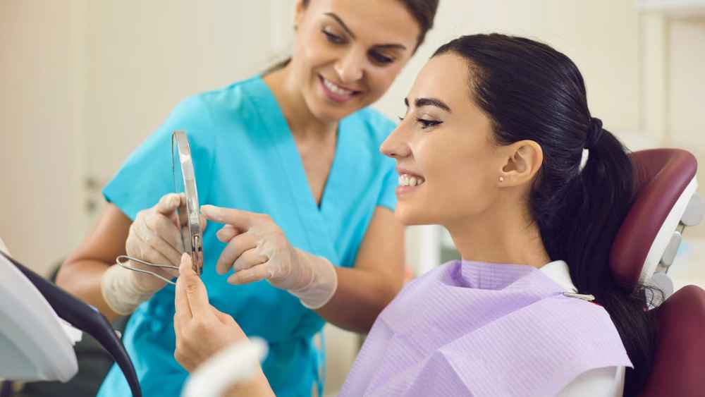 A young woman examines her teeth in the mirror of a dentists chair, admiring her smile post-treatment. The dental nurse is smiling beside her.
