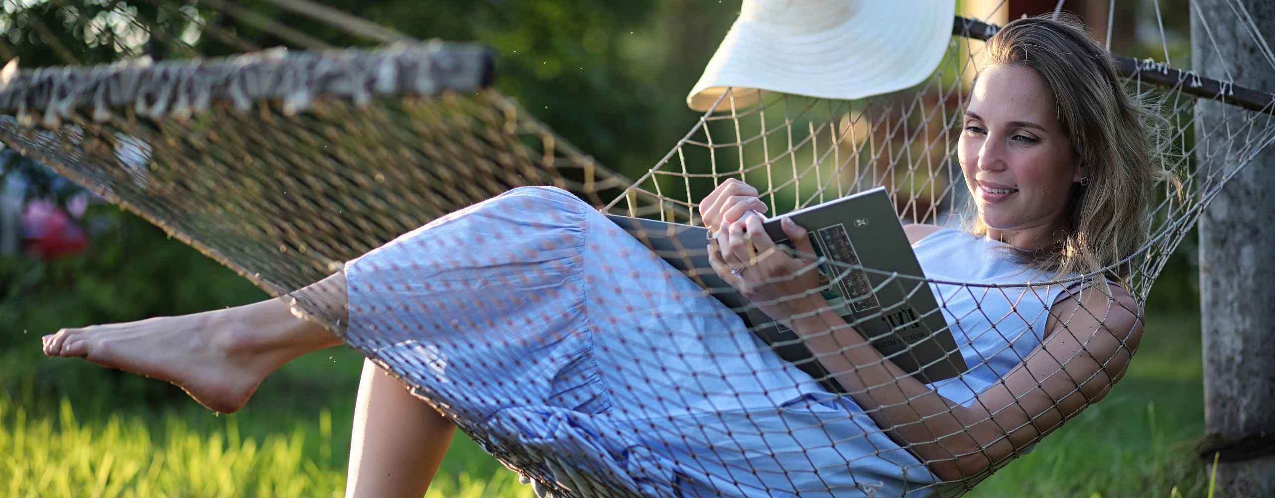 A smiling woman relaxing in an outdoor rope hammock while reading a book.