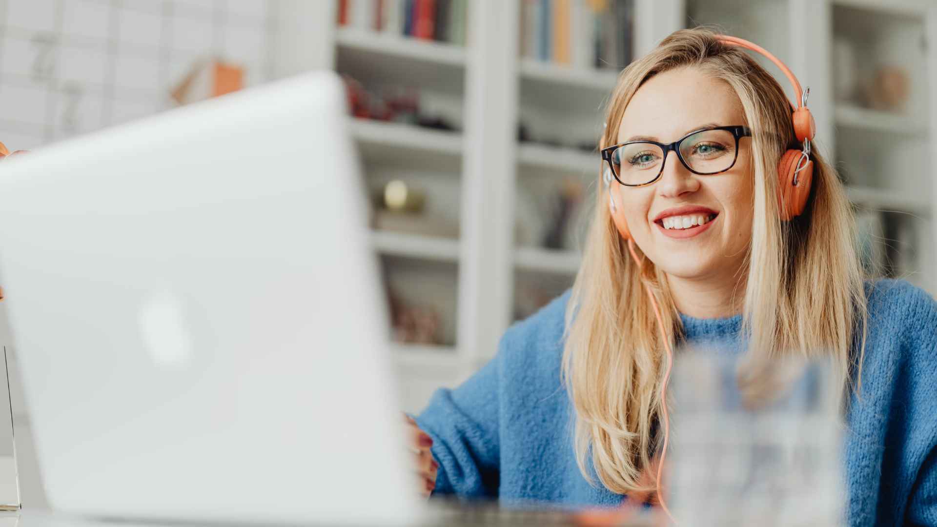 A woman in her 30's sits in her livingroom wearing glasses and earphones. She is smiling with her laptop open during a video call.