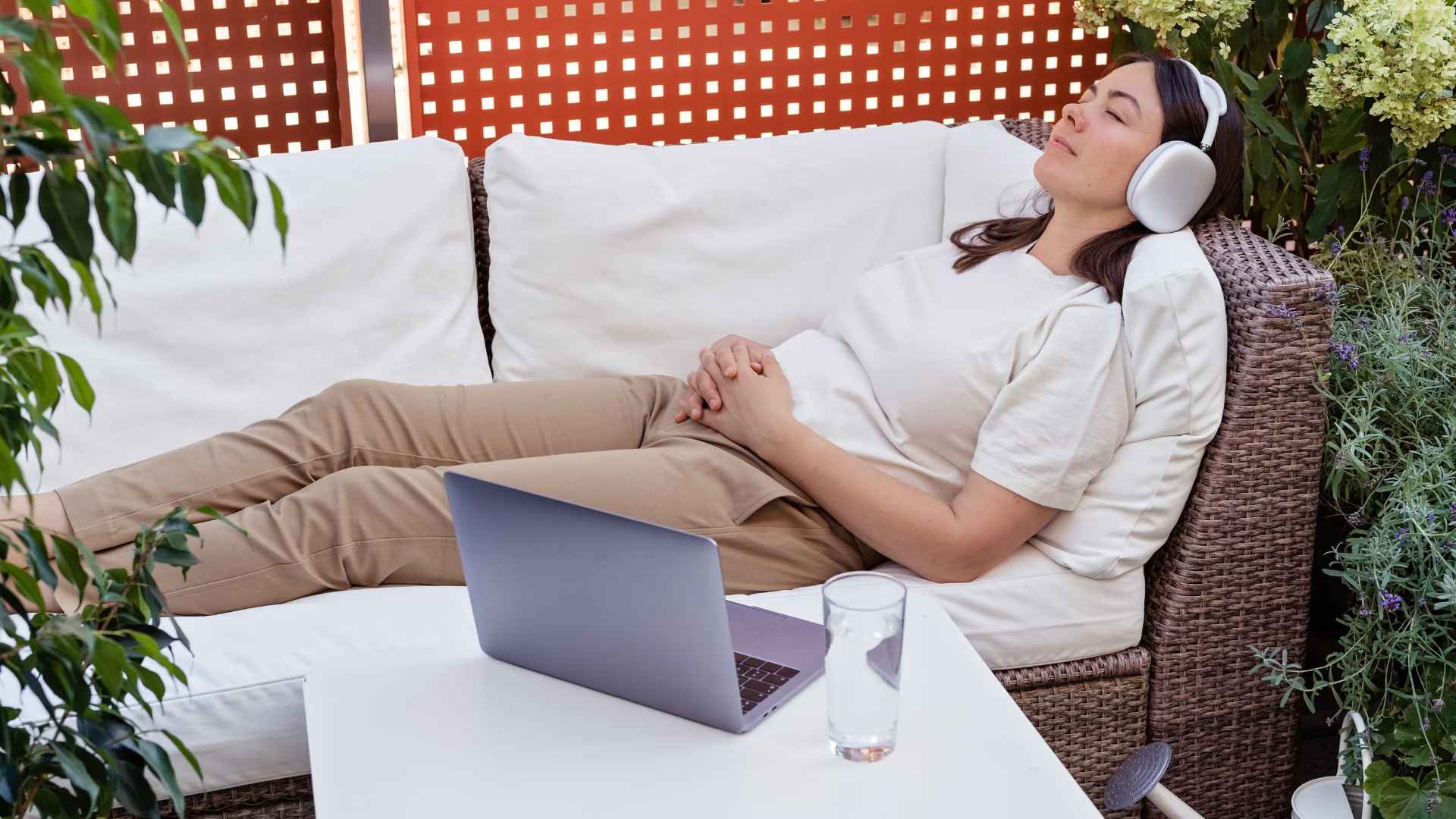 A woman reclining with her eyes closed on an outdoor couch, surrounded by greenery. She is wearing headphones with her laptop open on the table next to her. 